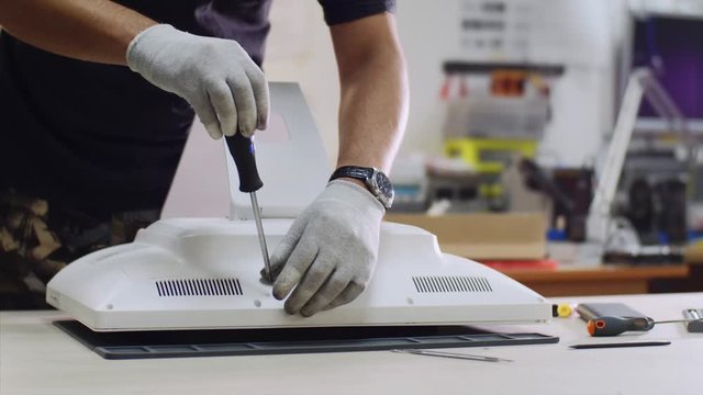 Repairman is working in electronics service center. Technician repairs and diagnostics computer monoblock unscrewing bolts with screwdriver in workshop on the table, hands in gloves closeup.