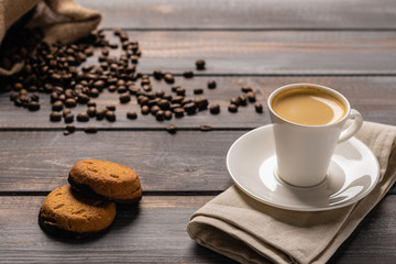 Cup of black coffee with cookies on a linen napkin on a dark wooden table. Coffee beans spill out of the bag on the table