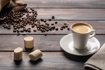 Cup of black coffee and a marshmallow on a linen napkin on a dark wooden table. Coffee beans spill out of the bag on the table