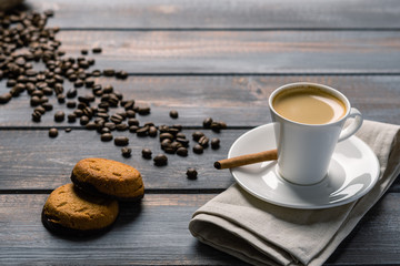 Cup of black coffee with cinnamon and cookies on a linen napkin on a dark wooden table. Coffee beans spill out of the bag on the table.