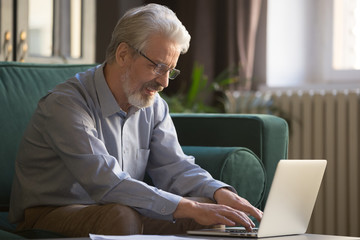 Smiling old man in glasses working on laptop at home
