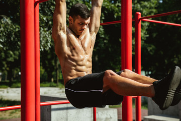 Young muscular shirtless caucasian man doing pull-ups on horizontal bar at playground in sunny summer's day. Training his upper body outdoors. Concept of sport, workout, healthy lifestyle, wellbeing.