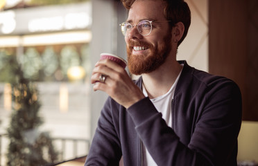 Joyful bearded man with ginger hair drinking coffee at cafe