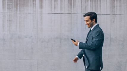 Happy Businessman is Walking with a Smartphone on a Street next to an Urban Concrete Wall. He's Wearing a Grey Suit. Sunny Day.