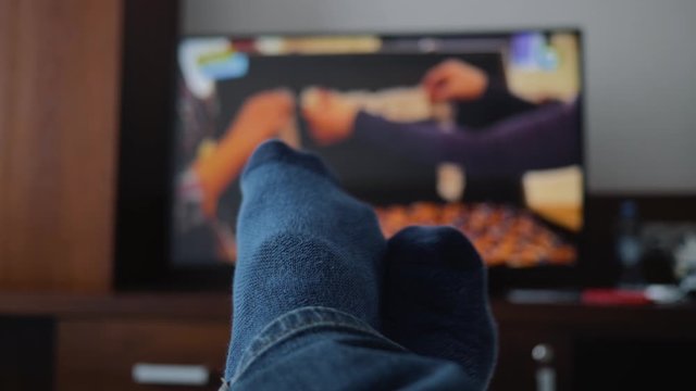 Legs in blue socks lie in front of the TV. Close-up on his feet. Cozy atmosphere of the room.