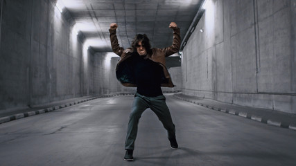 Fototapeta na wymiar Serious Cool Young Hipster Man with Long Hair is Energetically Dancing Hip Hop in a Lit Concrete Tunnel. He's Wearing a Brown Leather Jacket.