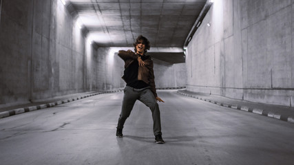 Serious Cool Young Hipster Man with Long Hair is Energetically Dancing Hip Hop in a Lit Concrete Tunnel. He's Wearing a Brown Leather Jacket.