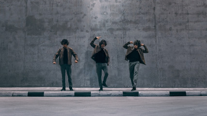 Multiple Exposure Shot of a Cool Cloned Young Hipster Man with Long Hair Stands in Dance Poses on a Street Next to a Big Concrete Wall. He's Wearing a Brown Leather Jacket.