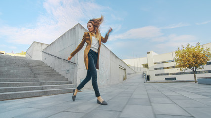 Cheerful and Happy Young Woman Actively Dancing While Walking Down the Stairs. She's Wearing a Long Brown Coat. Scene Shot in an Urban Concrete Park Next to Business Center. Day is Bright.