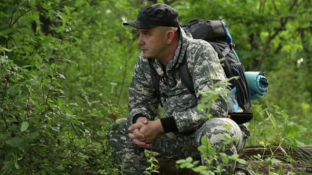 a young handsome caucasian man in a camouflage suit is resting on a log in a thick beautiful green forest and enjoying peace and tranquility, average plan