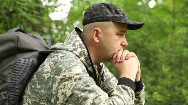a young handsome caucasian man in a camouflage suit is resting on a log in a thick beautiful green forest and enjoying peace and tranquility, average plan