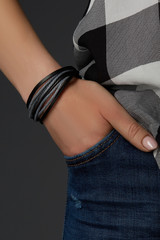 Cropped close-up shot of a female wrist with a multilayer bracelet with strings. The double-colored bracelet is made of gray wax ropes and black leather strips. The fashion model is dressed in jeans 