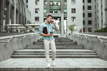 young handsome office worker talking on phone holding laptop and cup of coffee walking down the stairs at business center