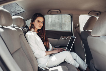 Fototapeta na wymiar Fashion Stylish Girl in White Suit Works at Laptop in Car
