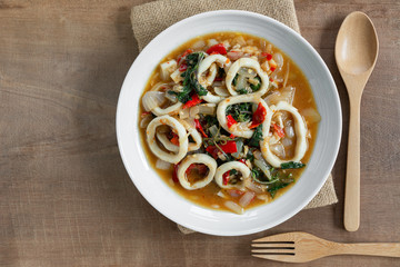 top view of stir fried squid with basil in a ceramic dish on wooden table. hot and spicy thai style food menu.