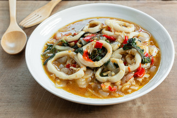 close up of stir fried squid with basil in a ceramic dish on wooden table. hot and spicy thai style food menu.