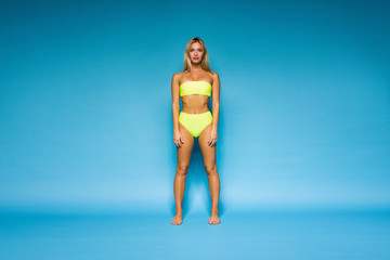 young tanned blonde girl in lemony yellow swimsuit is standing like fitness model and flirting with hands along the body on the blue wall background, sport lifestyle concept, free space