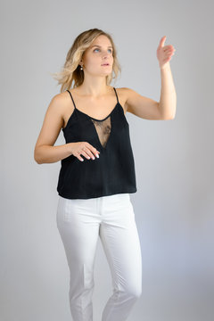 Full length studio portrait photo of a cute young blonde woman girl in a black blouse and white pants on a white background. He stands right in front of the camera, explains with emotion.