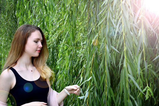 Young woman standing next to green willow tree under sun rays