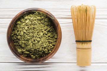 top view of wooden bowl with powder of green matcha tea on white table with whisk