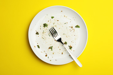 Dirty plate with food leftovers and fork on yellow background, top view