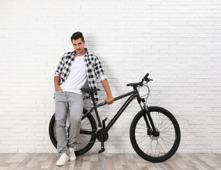 Handsome young man with modern bicycle near white brick wall indoors