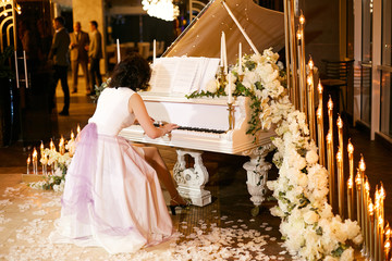 A girl in a beautiful dress plays the white piano during a wedding ceremony. Piano decorated with...