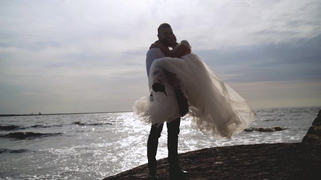 Groom holds bride in his arms on background of sea. Action. Groom picked up bride and turns her in sun on background of sea landscape. Sincere and radiant love of newlyweds