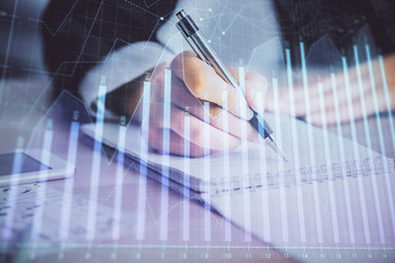 A woman hands writing information about stock market in notepad. Forex chart holograms in front....