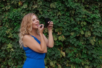 Beautiful young blonde girl in a dress, paints her lips with a small mirror. Real life, enjoy the moments. Copy space.