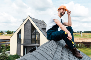 happy handyman in orange helmet sitting on roof and holding hammer