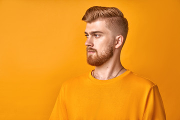 Fototapeta premium Profile shot of handsome male with trendy hairdo and red beard, looks aside with serious expression focused into distance, with blank space aside for your text or promotion, isolated yellow background