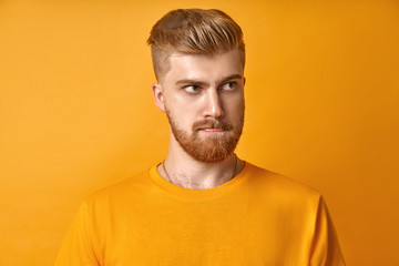 Puzzled handsome male with mustache and red beard, bites lower lip and looks curiously aside, thinks about something, dressed in casual yellow t shirt, stands against yellow background