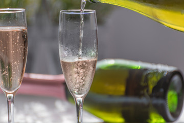 Close-up of a bottle pouring champagne into a glass, next to it a glass full of champagne and in the background a green bottle of unfocused champagne.