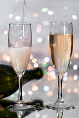 Serving sparkling champagne in a glass cup next to a glass with champagne and a green bottle behind with the background illuminated by many small lights.