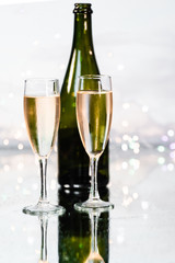 Two luxurious glass cups filled with yellow champagne and a green bottle behind with the background illuminated by many small Christmas lights. Concept of party and celebration.