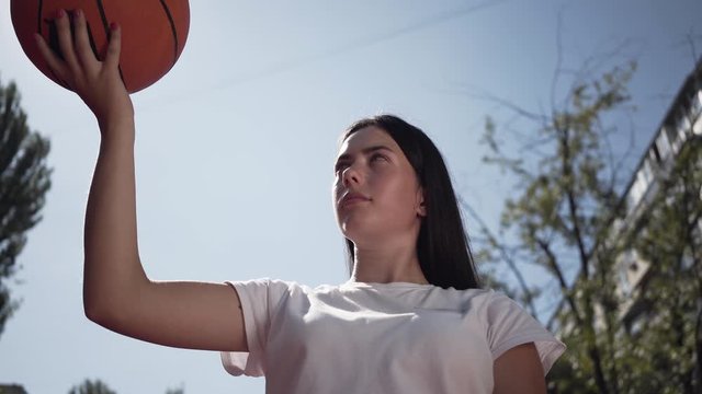 Portrait of cute young brunette woman throwing the ball up. Concept of sport, power, competition, active lifestyle. The girl playing basketball on the court. Bottom view. Sports and recreation.