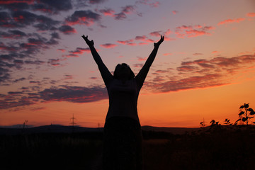 dark silhouette of a woman in a long dress that raised both hands up to the sky with a beautiful orange, pink sunset with purple clouds