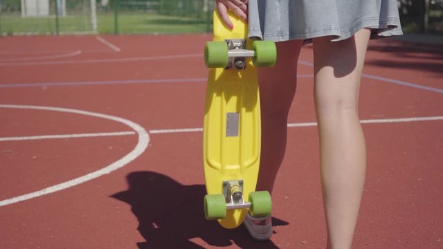 Legs of woman in short skirt with a yellow skateboard walking on the basketball court. Shadow follows the girl. Concept of sport, power, competition, active lifestyle