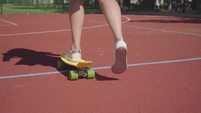Legs woman in skirt with a yellow skateboard walking on the basketball court. Female legs in sneakers girl riding yellow skateboard. Shadow follows the girl. Concept of sport, active lifestyle