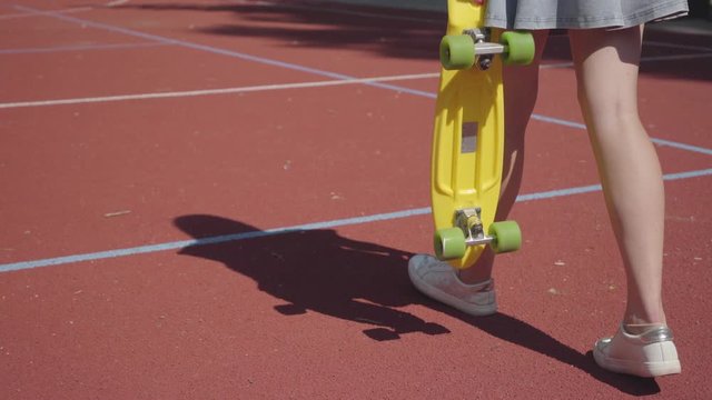 Legs woman in short skirt with a yellow skateboard walking on the basketball court. Female legs in sneakers girl riding yellow skateboard. Shadow follows the girl. Concept of sport, active lifestyle