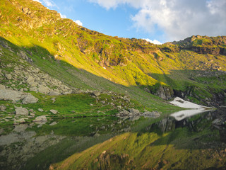 Beautiful landscape with high mountains with illuminated peaks, stones in mountain lake, reflection, blue sky and yellow sunlight in sunrise.