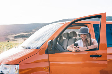 pretty and attractive blonde girl with cap wearing phone in orange van with mountains in the background and gray sky