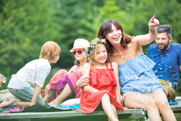 Smiling woman pointing to daughter while sitting on pier against family at lakeshore during summer