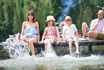 Photo of mother playing water with kids in lake while sitting on pier against family at lakeshore during summer - obrazy, fototapety, plakaty