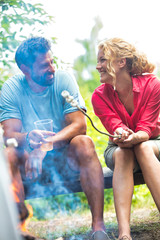 Smiling man drinking beer and a woman roasting marshmallows over burning campfire at park