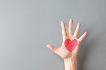 Hand palm of young Caucasian woman girl with painted on red heart on gray wall background. Charity...