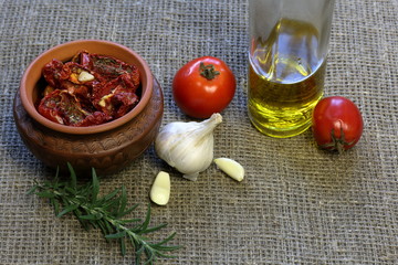 Sun-dried tomatoes with spices and garlic in a clay pot. Nearby is a bottle with olive oil, tomatoes, rosemary and garlic.
