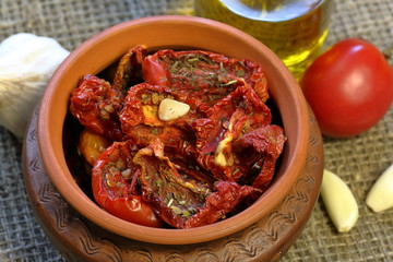Sun-dried tomatoes with spices and garlic in a clay pot. Nearby is a bottle with olive oil, tomatoes, rosemary and garlic.
