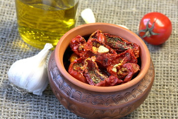 Sun-dried tomatoes with spices and garlic in a clay pot. Nearby is a bottle with olive oil, tomatoes and garlic.